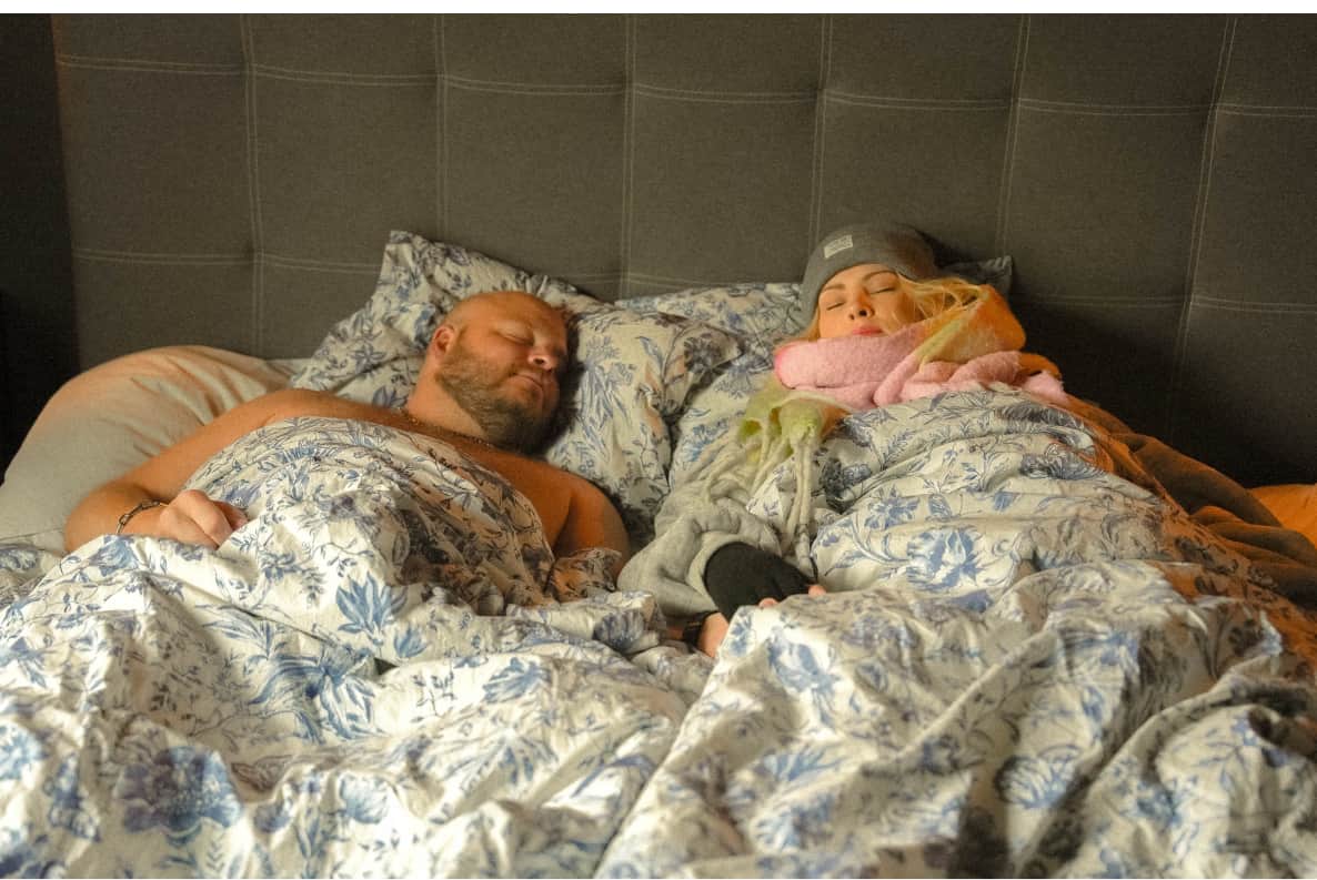 Mirja and Jimmi asleep. She is in a beanie and scarf under blankets, he has no duvet.