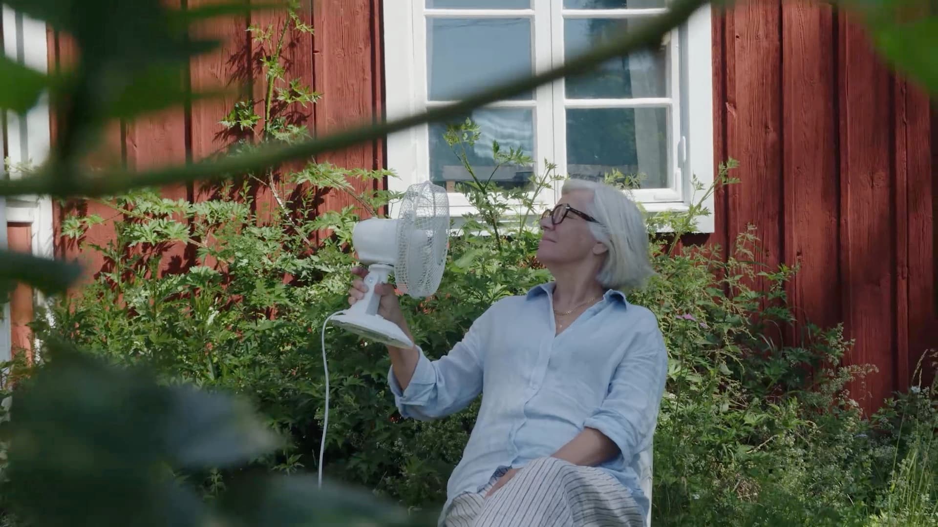 Older Elvy customer cooling off with a desk fan outside a red house