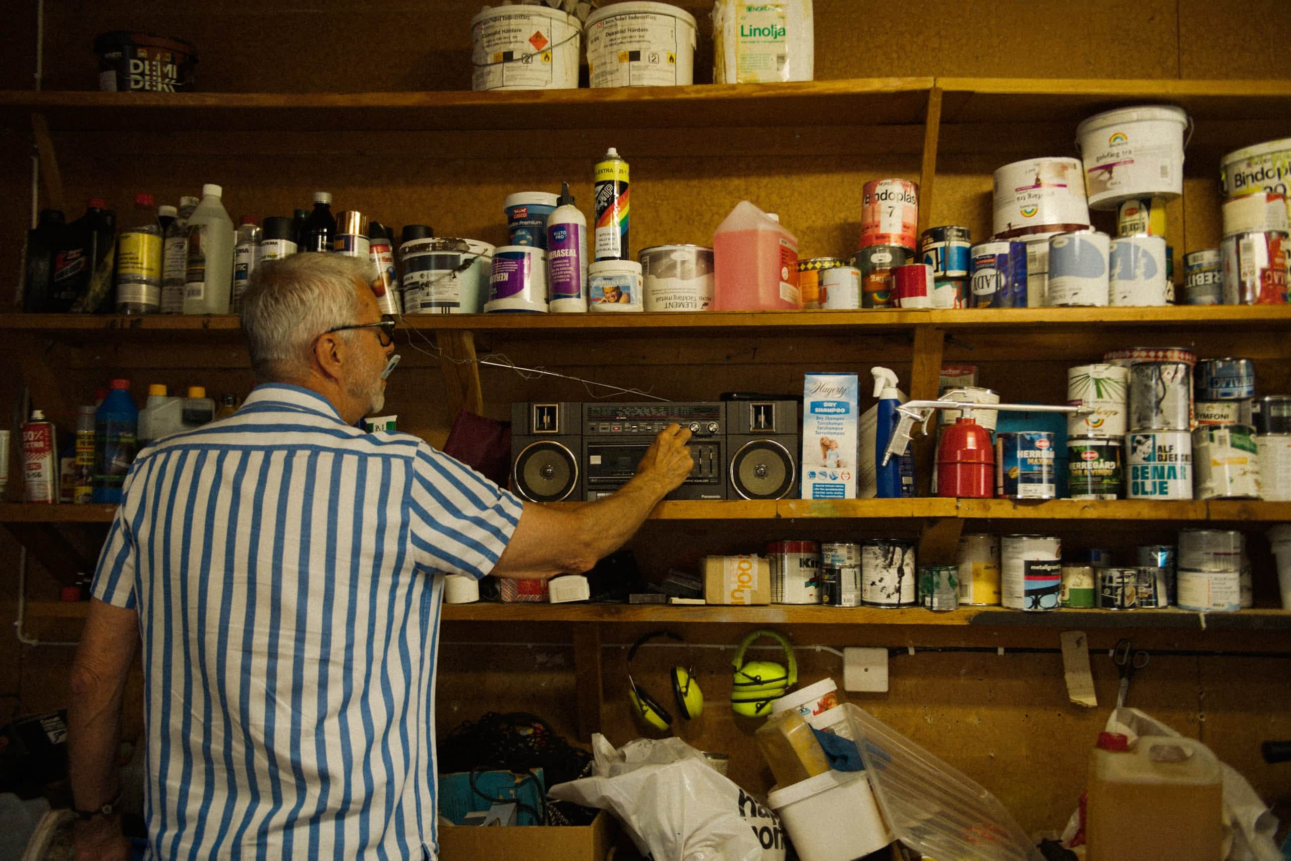 Older Elvy customer reaching for an old radio among paint-can shelves in a farm storage room